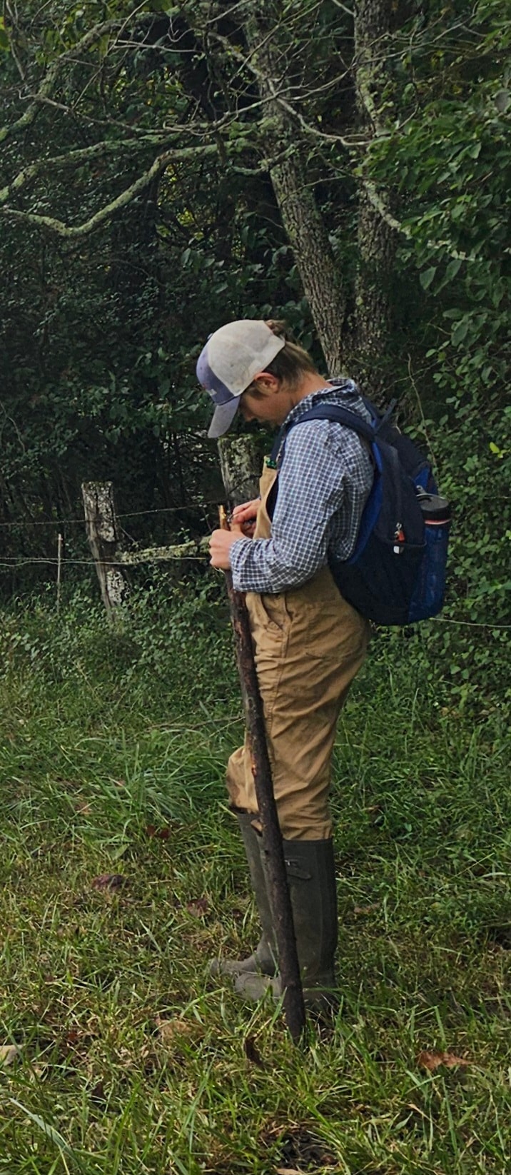 Student in overalls and rubber boots leaning on a walking stick at the forest edge